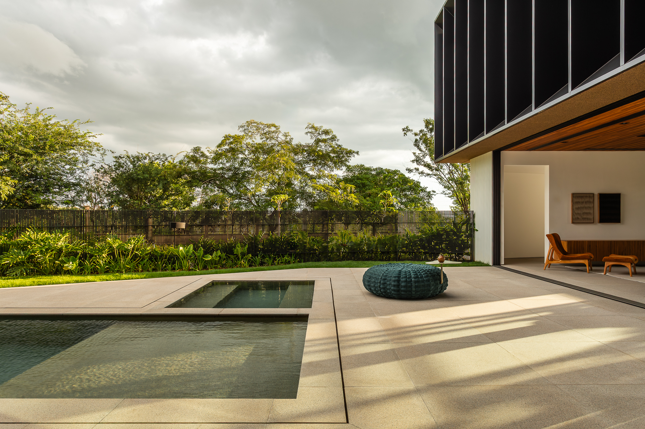 Casa Buritis - Área externa com piscina geométrica, puff trançado verde-azulado e vista da integração interior-exterior ao entardecer