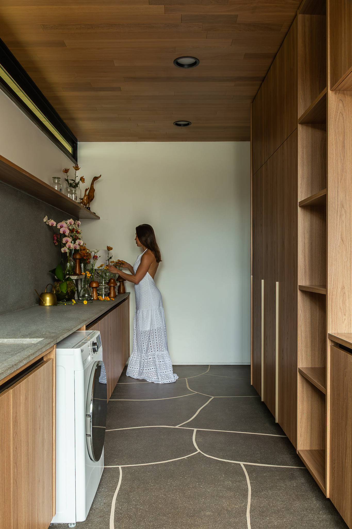 Casa Funchal - Lavanderia com teto de madeira, armários personalizados, piso com padrão orgânico e arranjos florais decorativos