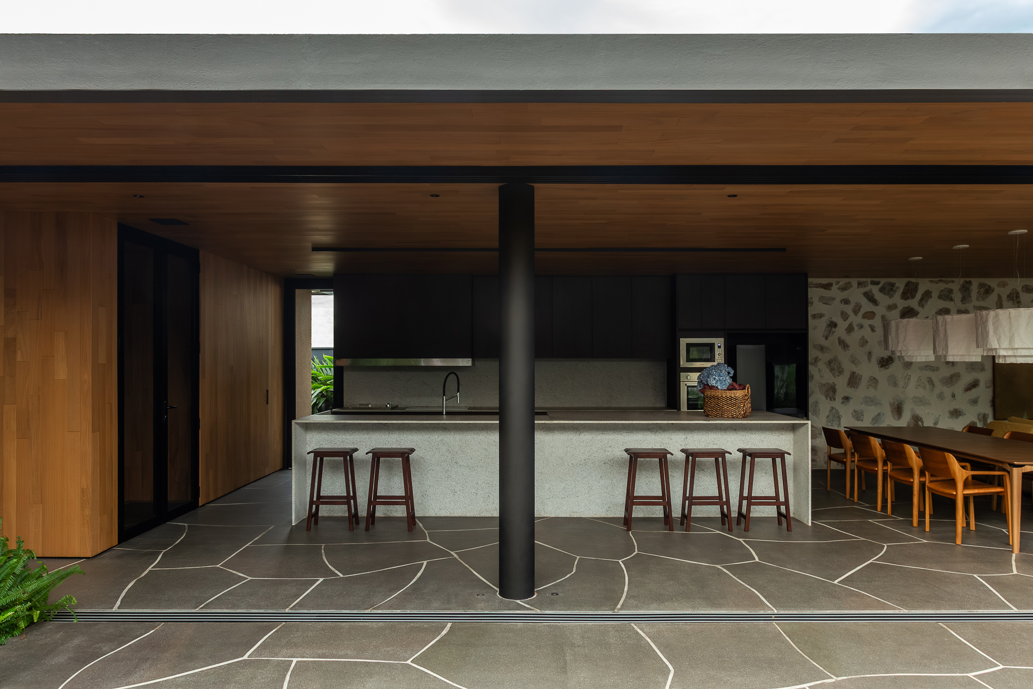Casa Funchal - Cozinha externa coberta com ilha de terrazzo, banquetas de madeira, coluna estrutural preta e continuidade do piso com padrão orgânico