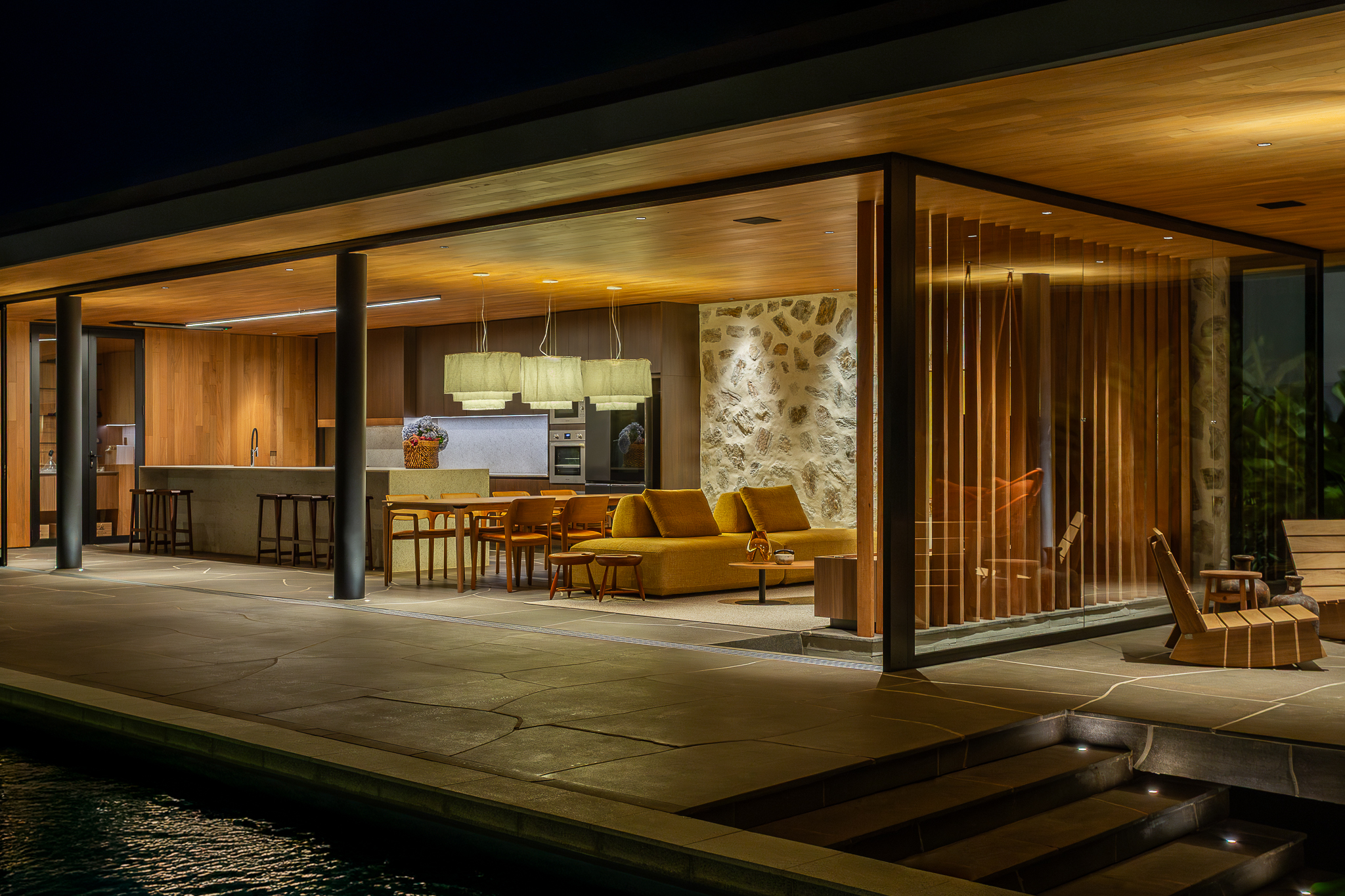 Casa Funchal - Vista noturna externa do pavilhão envidraçado com teto de madeira, sofá amarelo mostarda, área de jantar e piscina em primeiro plano