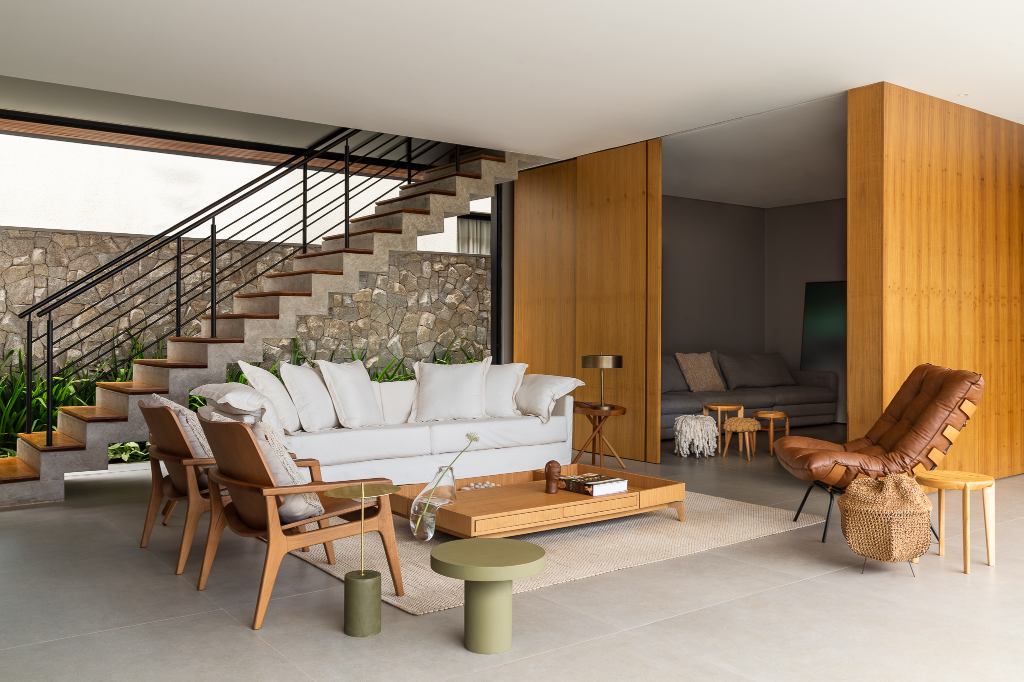 Casa Bosque - Sala de estar principal com pé-direito duplo, escada em pedra, sofá branco, poltronas de madeira e divisória em madeira