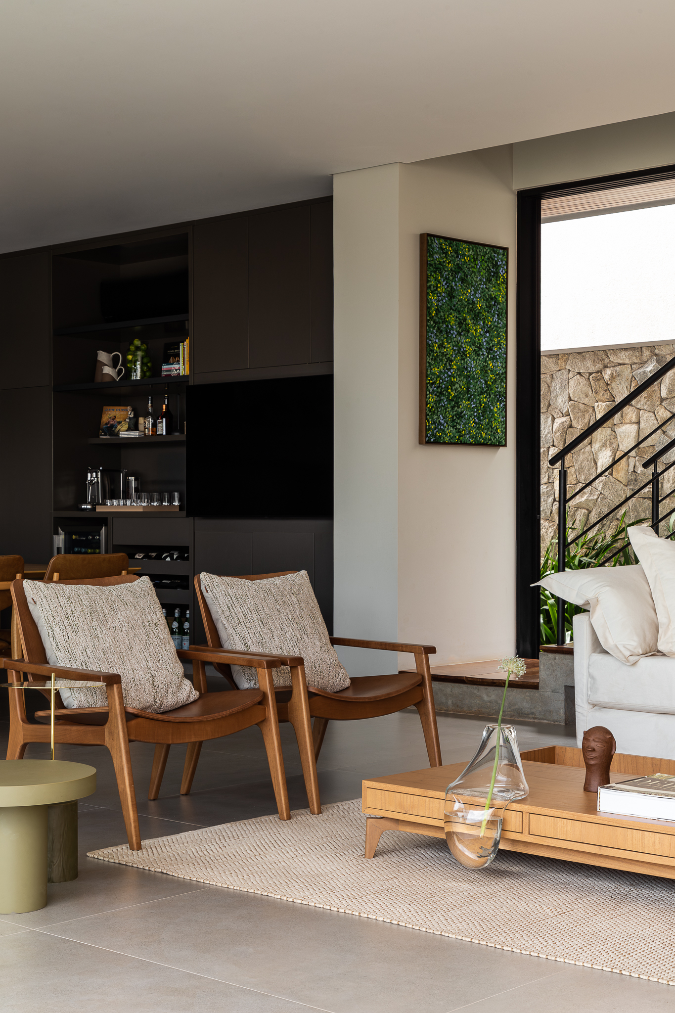 Casa Bosque - Sala de estar com poltronas de madeira, obra têxtil verde-azulada e vista para área externa com pedras