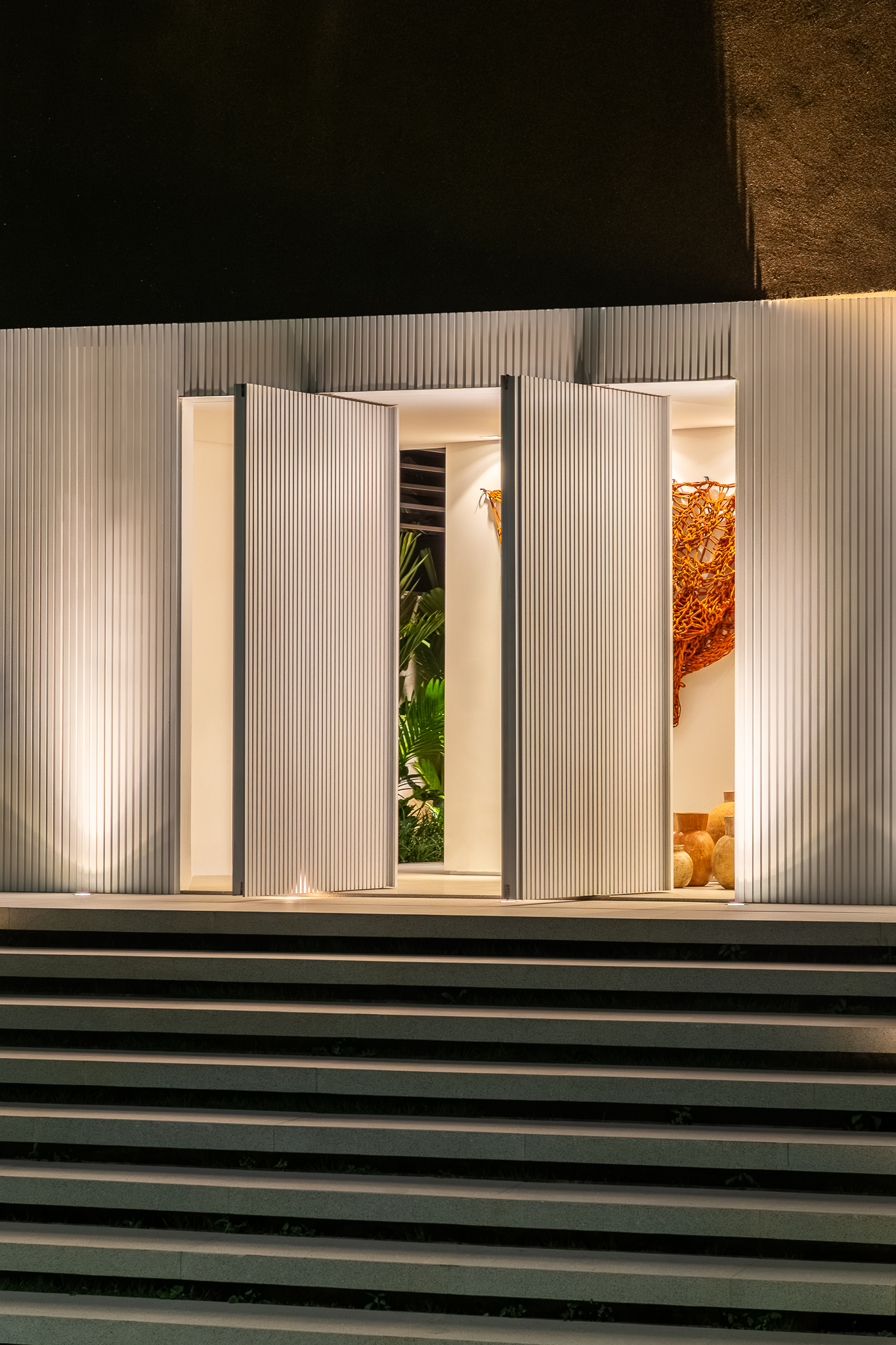 Casa Buritis - Detalhe noturno da entrada com fachada ripada iluminada, obra têxtil laranja queimado em destaque e escada em primeiro plano