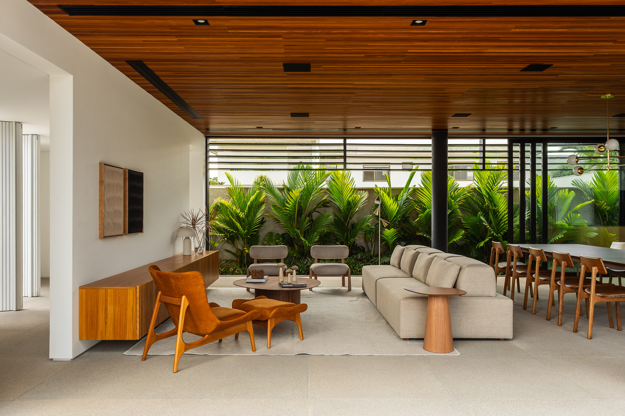 Casa Buritis - Sala de estar e jantar integradas com teto ripado em madeira, poltrona laranja queimado, sofá bege modular e vista para jardim tropical