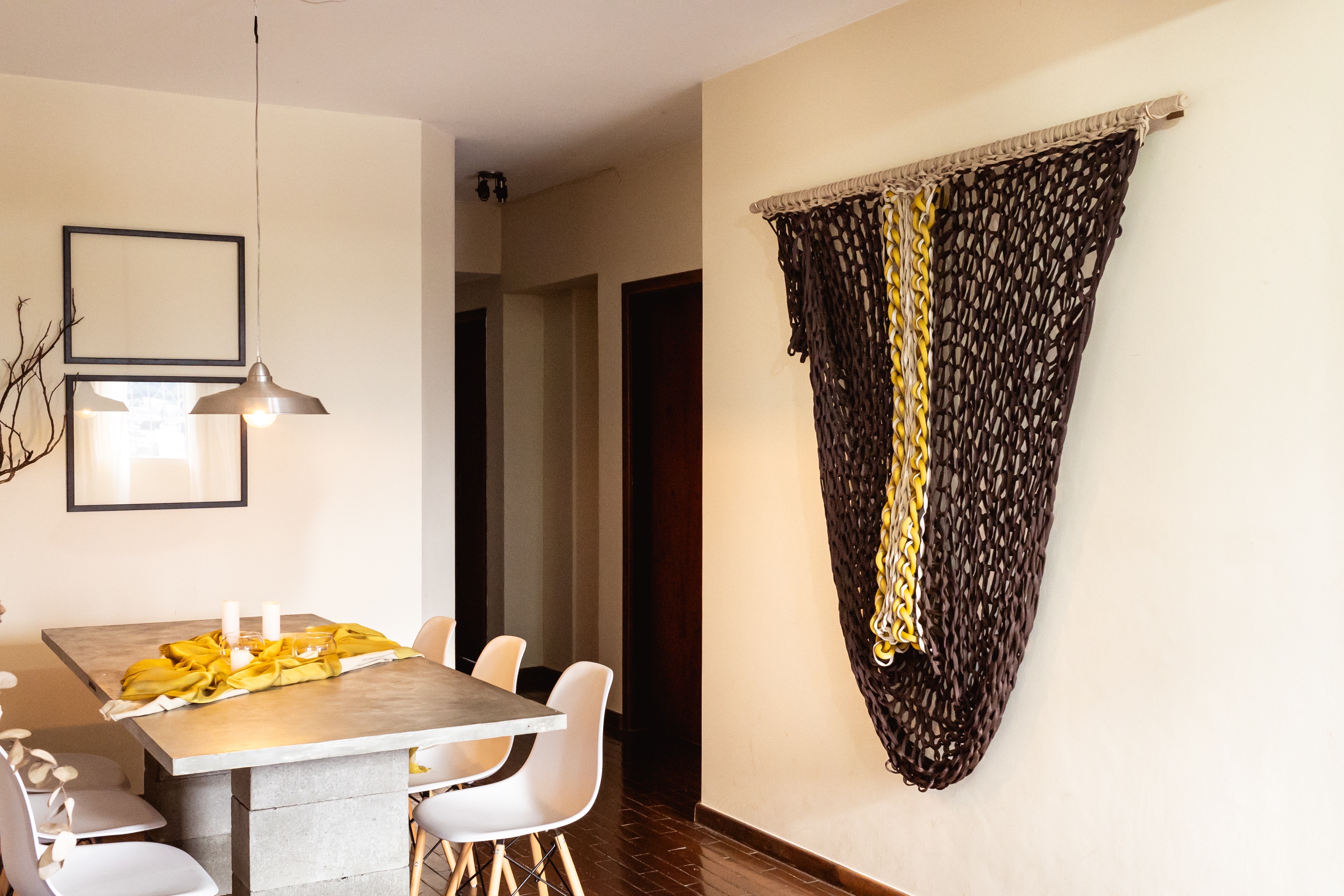 Escultura têxtil triangular em malha de macramé marrom escuro com faixa vertical amarela, instalada em ambiente de jantar