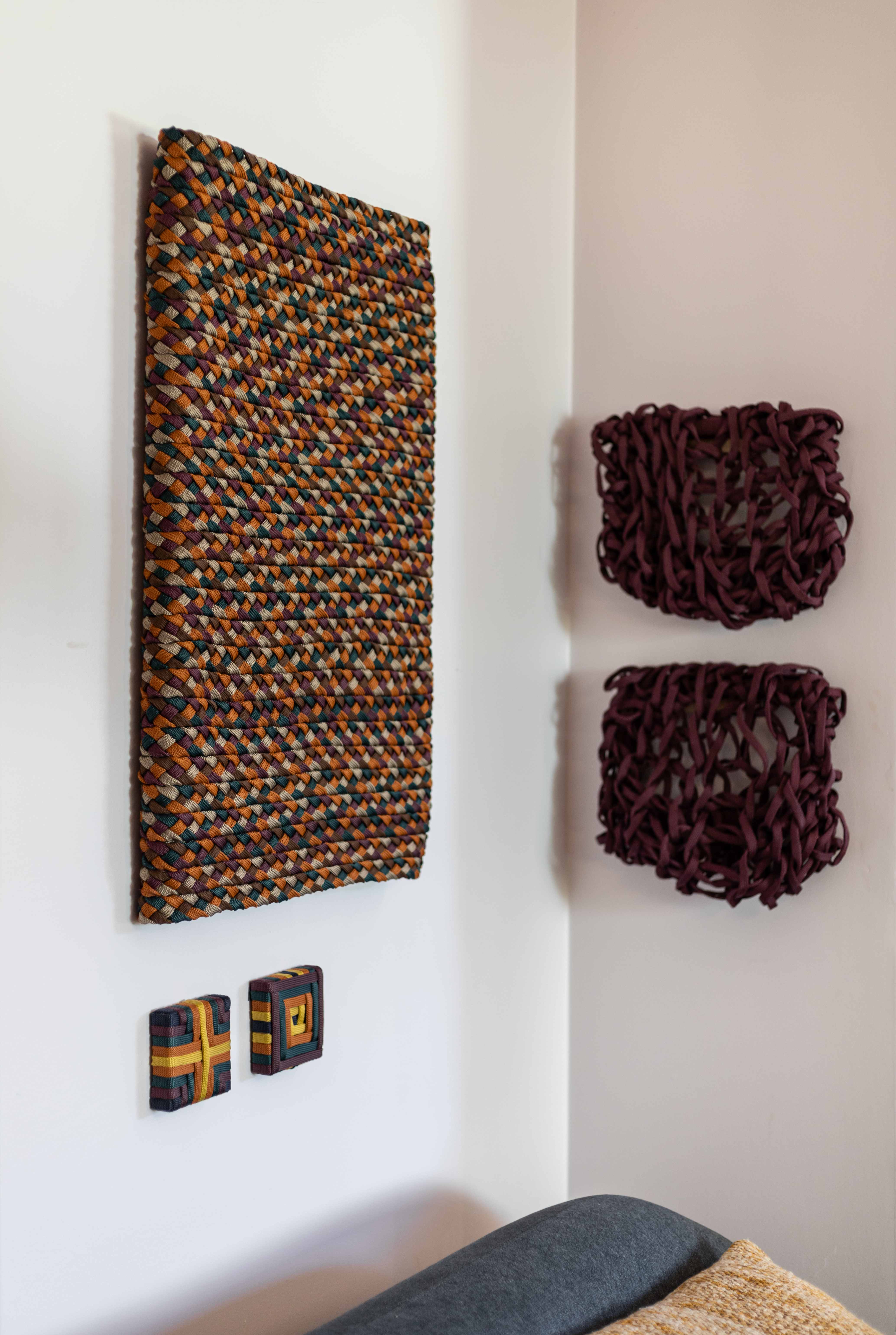 Composição de parede com peças têxteis tecidas em diversas escalas e cores, incluindo padrões multicoloridos e esculturas em tons de vinho